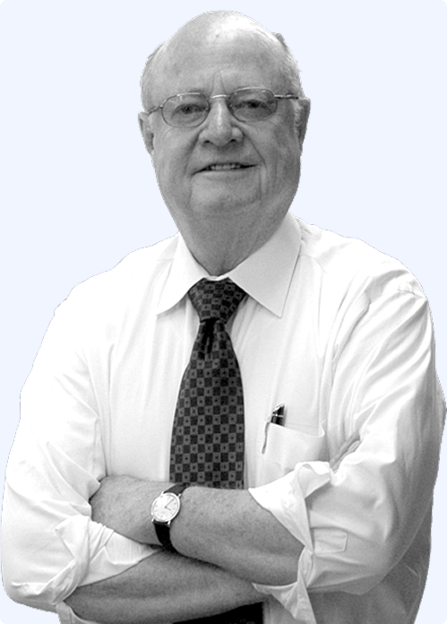 An older man with glasses wearing a white shirt and a patterned tie, posing with his arms crossed.