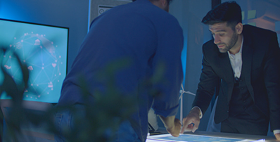 Two men in a dimly lit office examine data on an illuminated touchscreen table, with a digital network graphic visible on a monitor in the background.