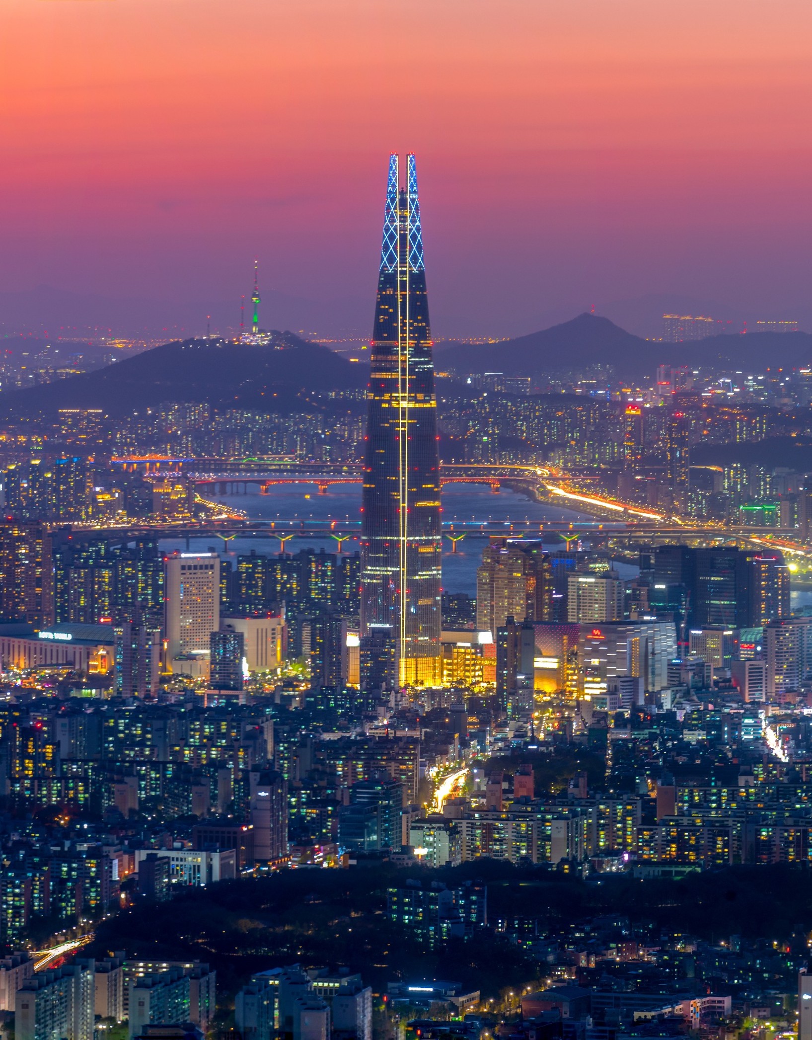 The Lotte World Tower stands prominently over the Seoul skyline at dusk, with the Han River and city lights glowing against a purple and orange sunset.