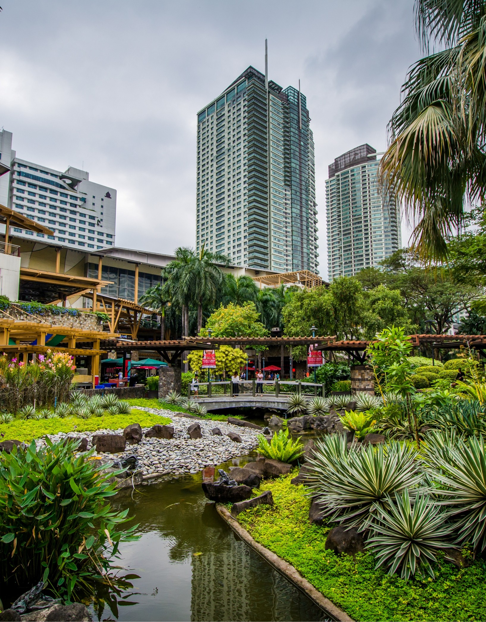 A landscaped garden with a small pond and stone-lined stream leading to a footbridge, set against a backdrop of modern high-rise buildings and lush trees.