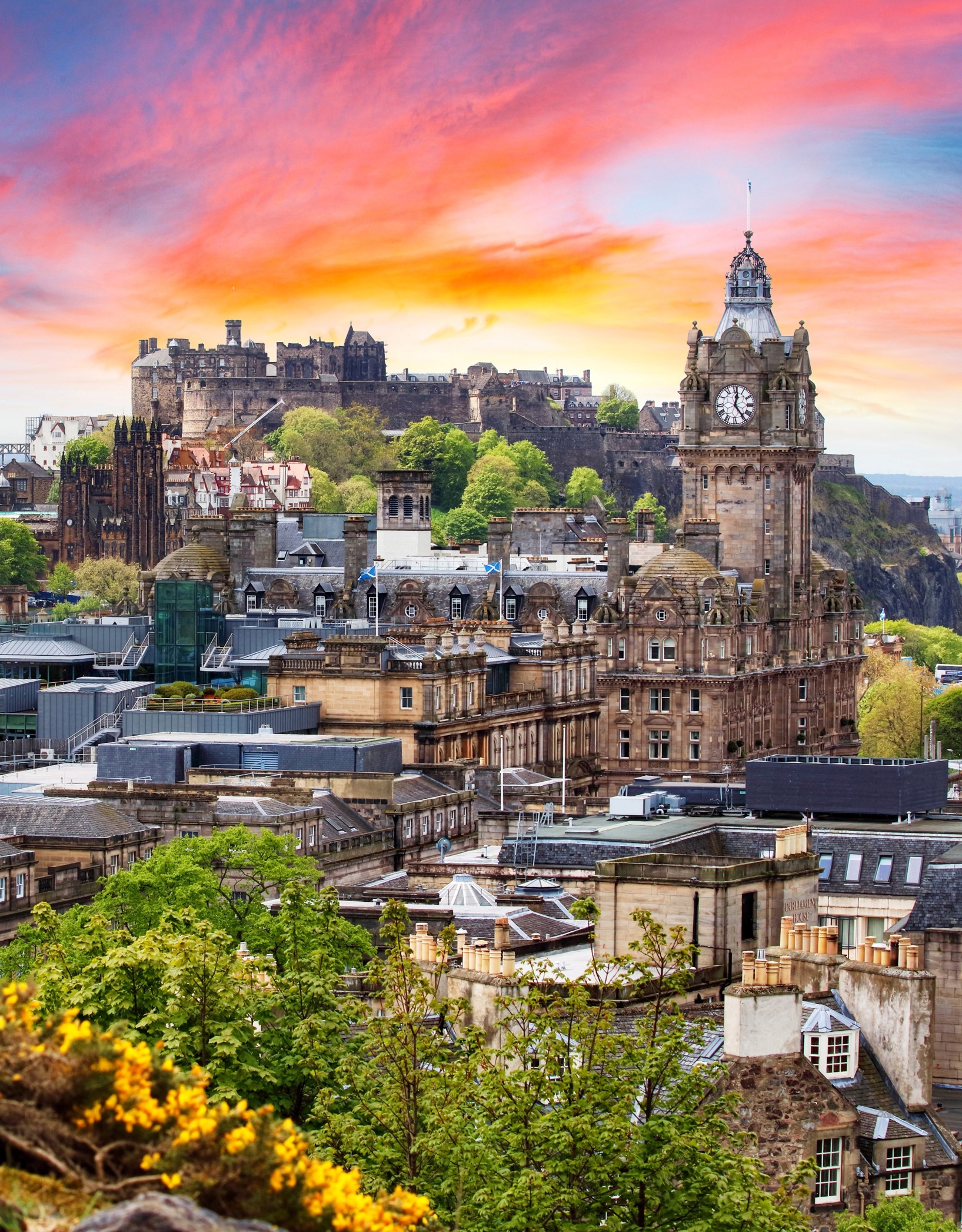 Edinburgh Castle overlooks the Balmoral Hotel clock tower beneath a vibrant orange and pink sunset.