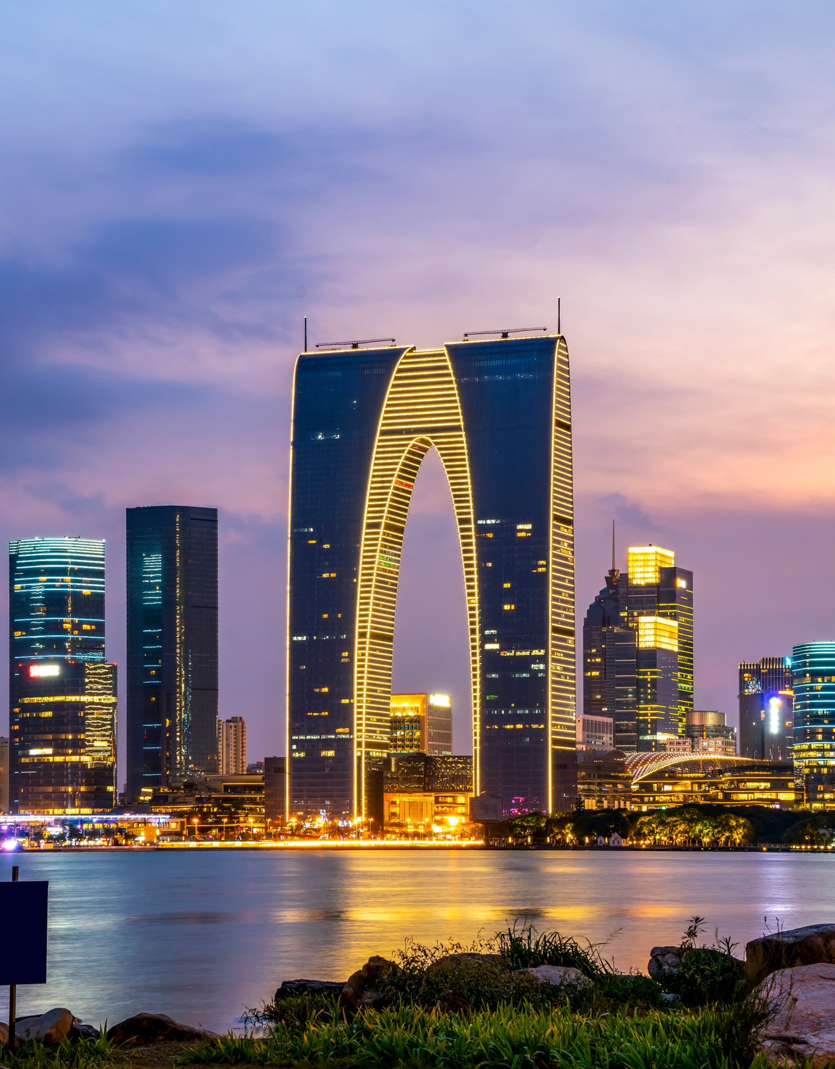 The Gate to the East skyscraper standing prominently among a city skyline at dusk, reflected in a body of water.
