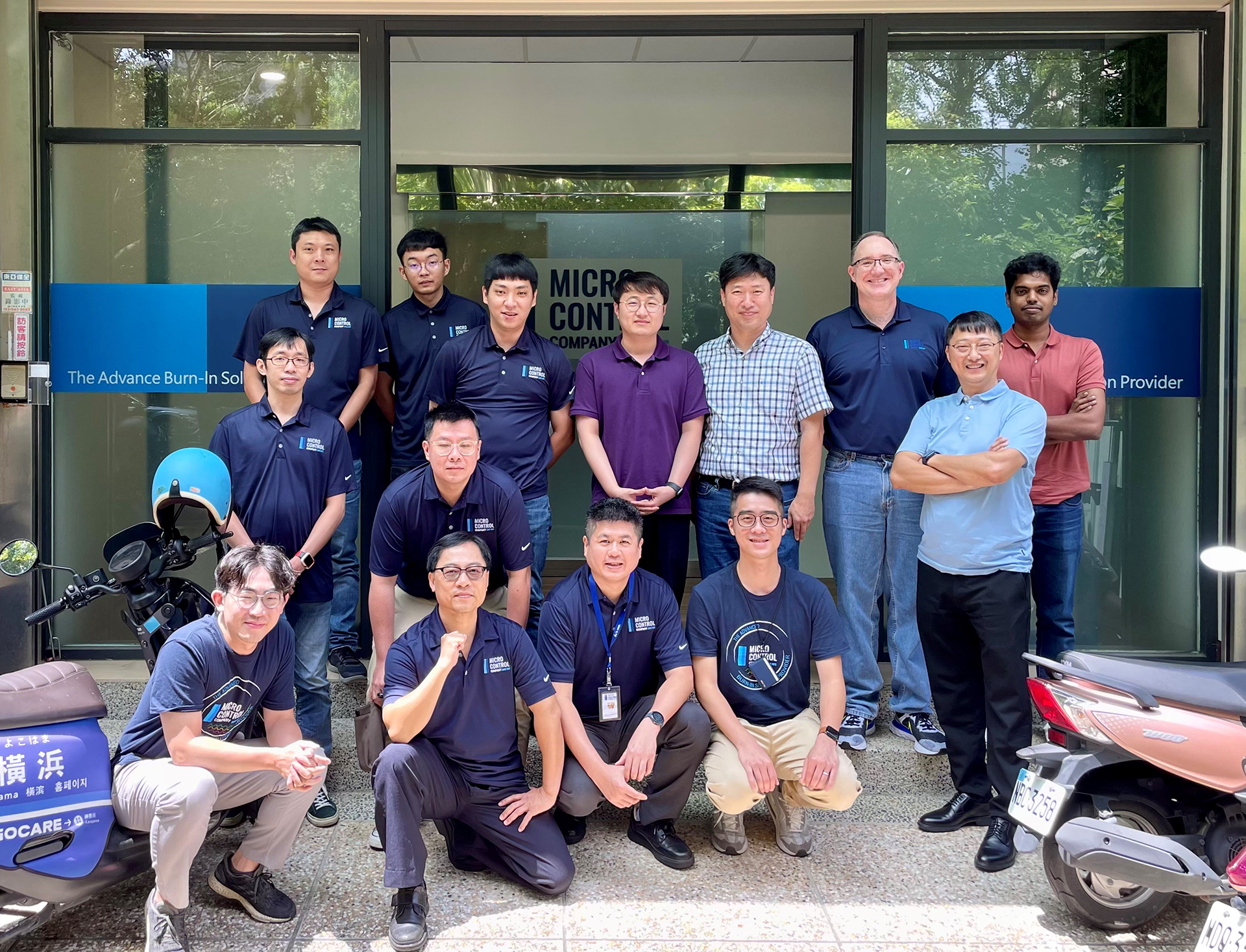 A group of fourteen people standing and crouching in front of the Micro Control Company office entrance.