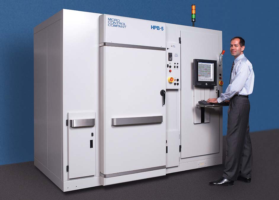 A man in a dress shirt and tie stands at the control panel of a large Micro Control Company HPB-5 industrial test chamber.