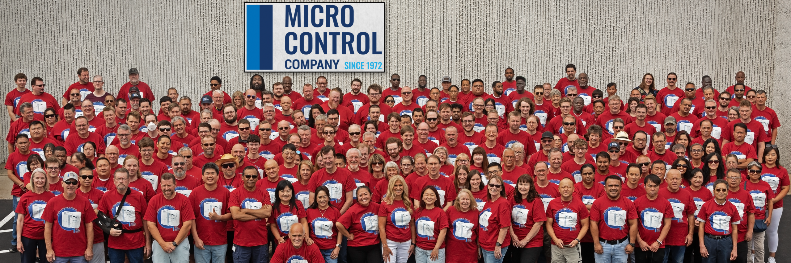 A large group of people posing for a photo in front of a white wall with the Micro Control Company logo.